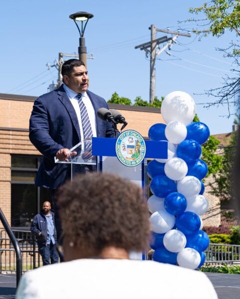 Alderman Chico speaks at the podium during the Saturday Morning Quantum graduation ceremony, standing beside a column of blue and white balloons and addressing students, families and community members.