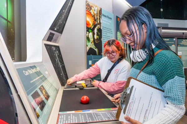 Two high school students participate in a hands-on Saturday Morning Quantum activity at Fermilab, engaging with a display titled “Can You Beat Light?”
