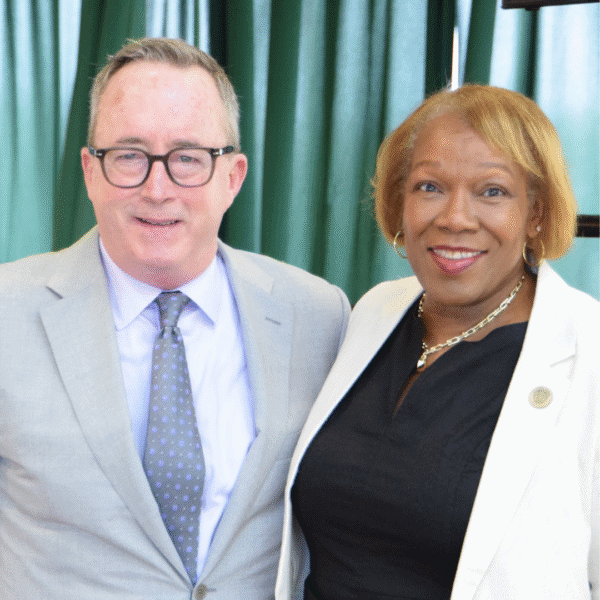 Two smiling individuals pose together at an event. John Atkinson, on the left, wears glasses and a light gray suit with a dotted tie. Z Scott, on the right, wears a white blazer over a black dress. They stand in front of green curtains.
