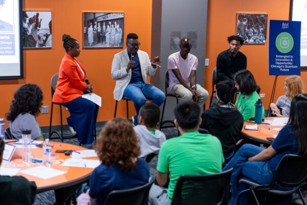 A panel of four speakers engages with an audience of students. One panelist speaks into a microphone while others listen, seated on stools in front of an orange wall with historic photos and an event sign reading “Entangled in Innovation & Opportunity: Chicago’s Quantum Future.”
