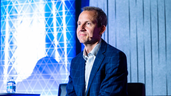 Dr. Harley Johnson, CEO of the Illinois Quantum and Microelectronics Park (IQMP), sits on stage wearing a blue checkered blazer and headset microphone, with a geometric backdrop behind him.