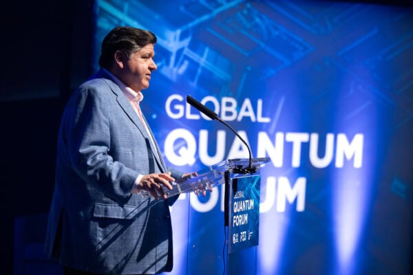 Governor JB Pritzker delivers opening remarks at the Global Quantum Forum in Chicago, standing at a clear podium in front of a blue-lit stage backdrop featuring the event’s name.