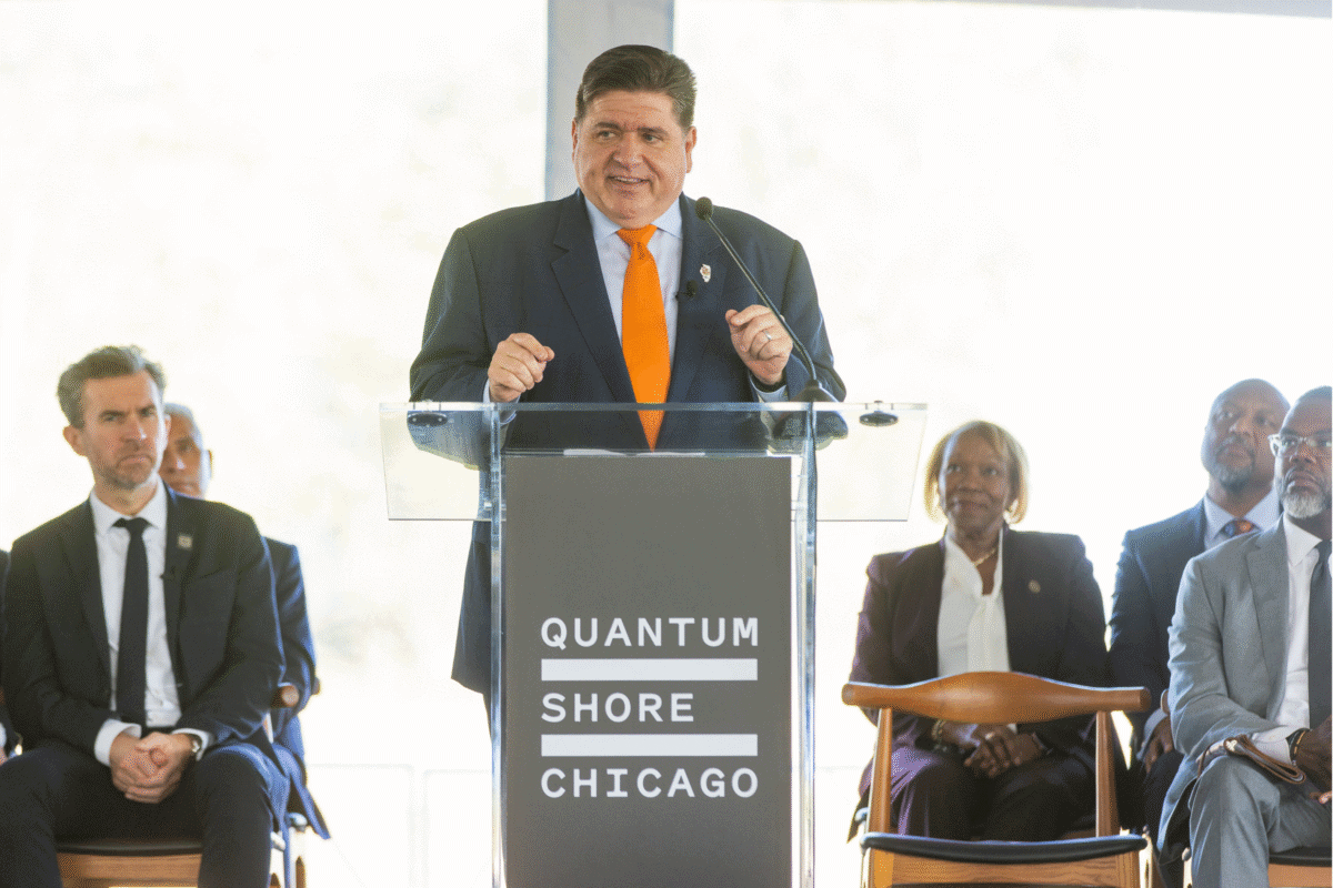Governor JB Pritzker, wearing a dark suit and orange tie, speaks at a podium labeled “Quantum Shore Chicago" during the groundbreaking event.