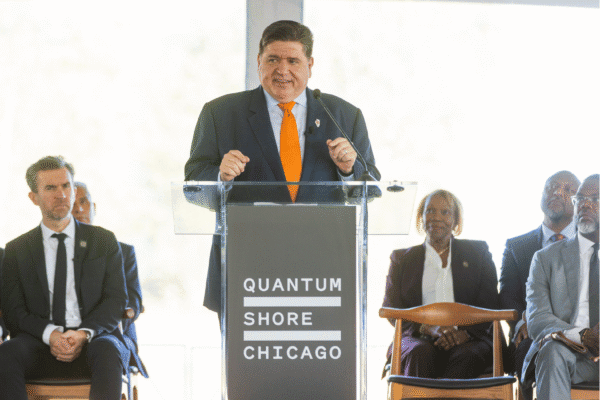 Governor JB Pritzker, wearing a dark suit and orange tie, speaks at a podium labeled “Quantum Shore Chicago" during the groundbreaking event.