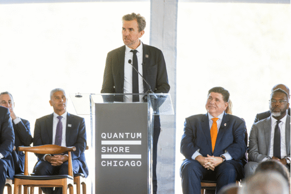 Dr. Jeremy O'Brien, wearing a dark suit and dark tie, speaks at a podium labeled “Quantum Shore Chicago" during the groundbreaking event.