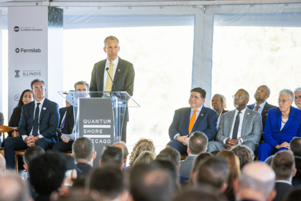 Dr. Harley Johnson, wearing a grey suit and yellow tie, speaks at a podium labeled “Quantum Shore Chicago" during the groundbreaking event.