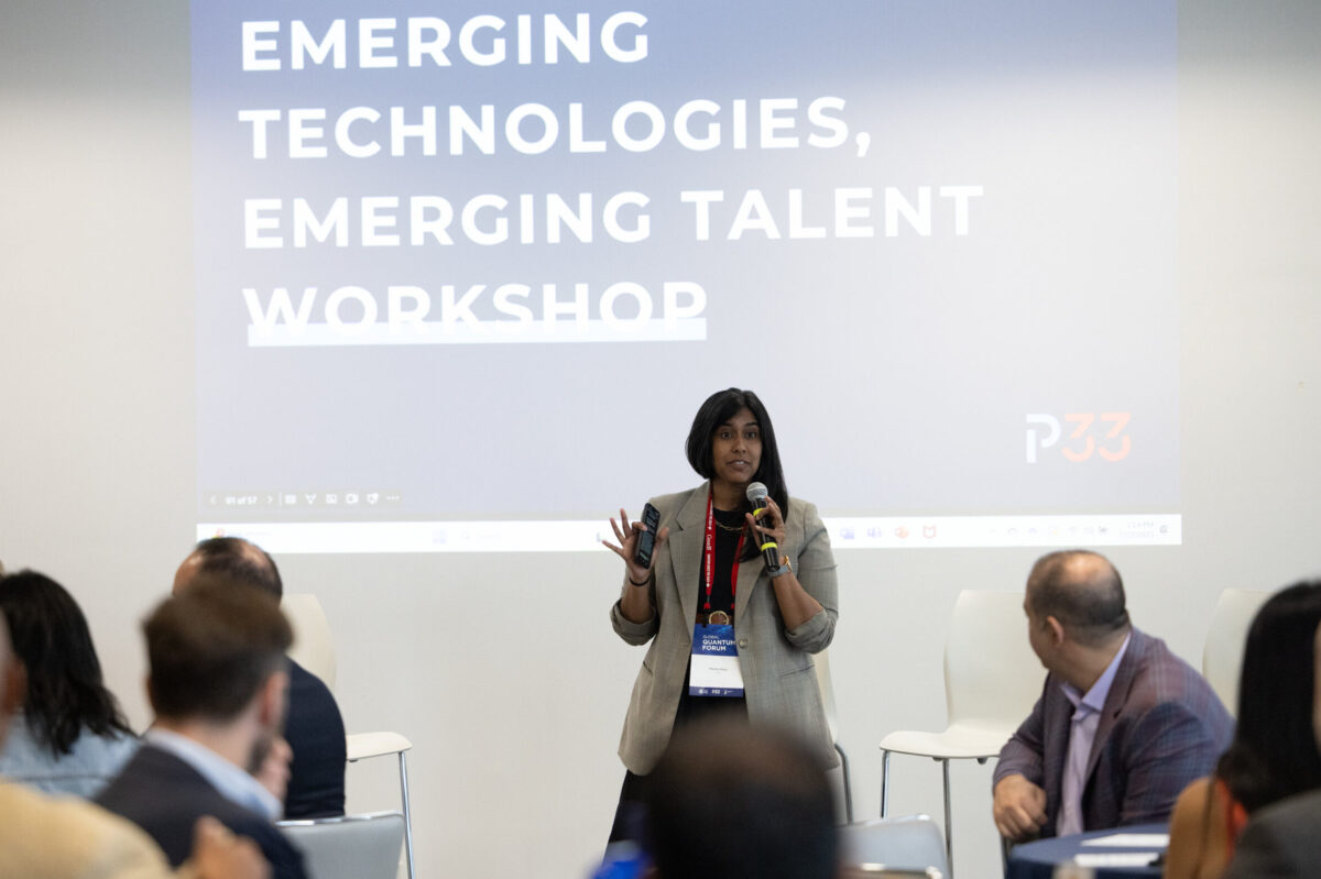 Meera Raja speaks to an audience at a workshop, holding a microphone and clicker, with a large screen behind her displaying the words “Emerging Technologies, Emerging Talent Workshop.”