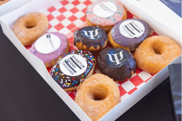 A box of decorated donuts sits on a table, featuring frosting designs with logos for “Quantum Shore Chicago” and PsiQuantum.